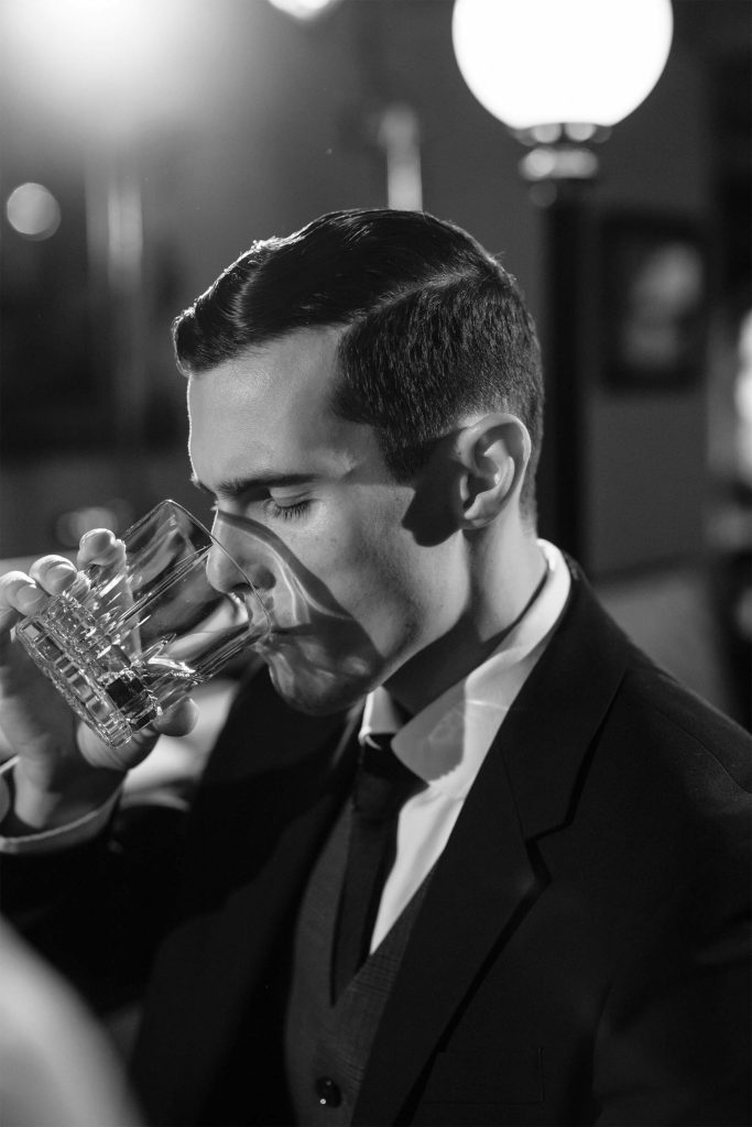 Schwarz-Weiß-Foto eines eleganten Mannes in einem Anzug, der aus einem Glas trinkt, mit unscharfen Lichtern im Hintergrund.